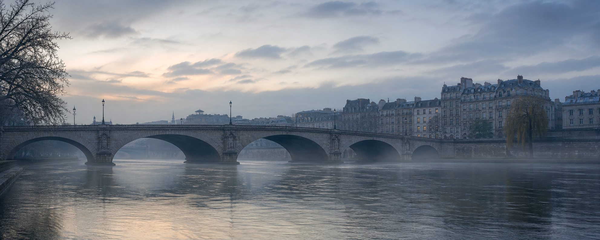 A stone arch bridge over a still river at dawn, with mist on the water and a line of stone buildings beyond.