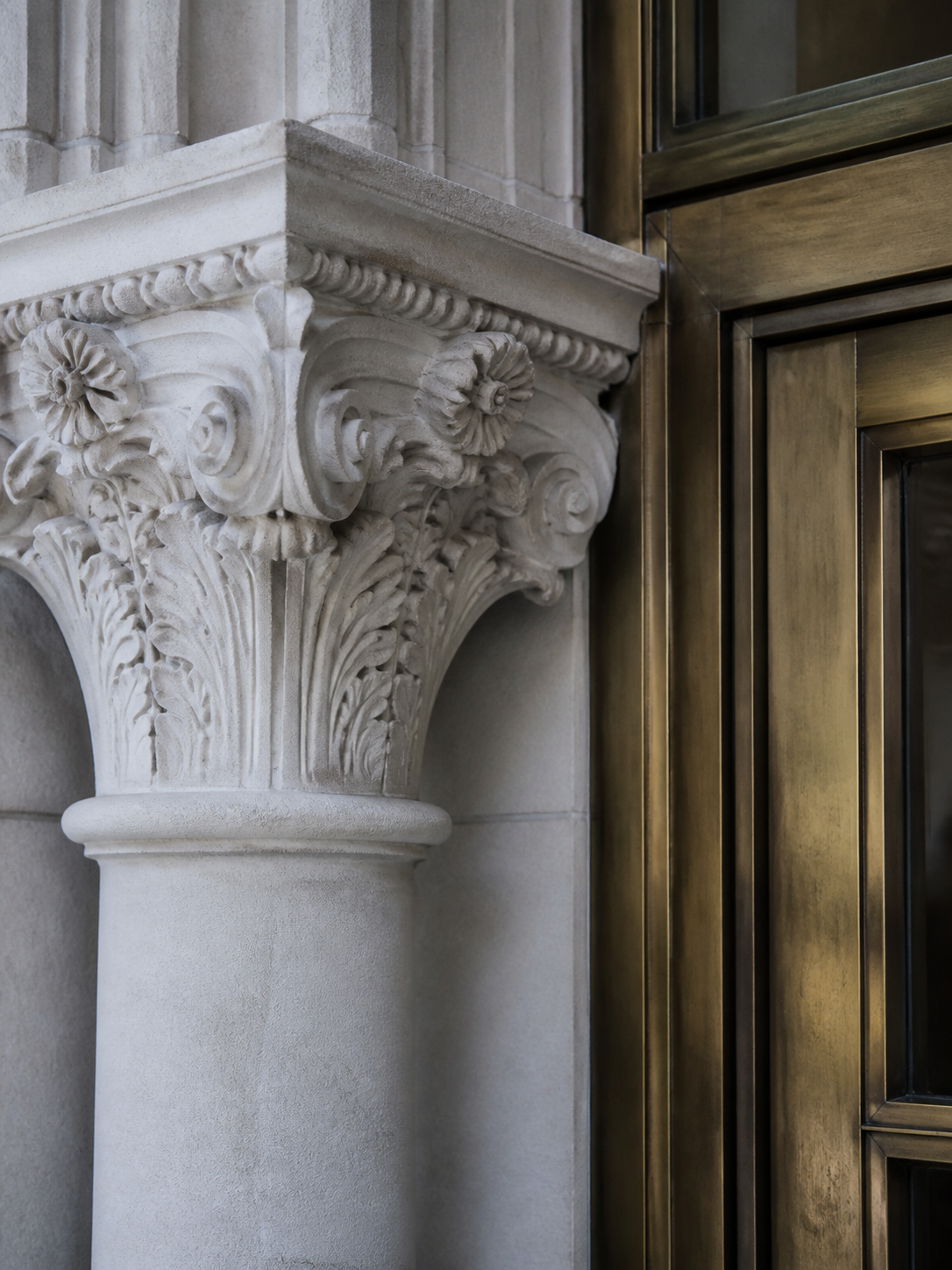 A carved stone column capital beside the tall brass frame of an entrance door, in cool even daylight.
