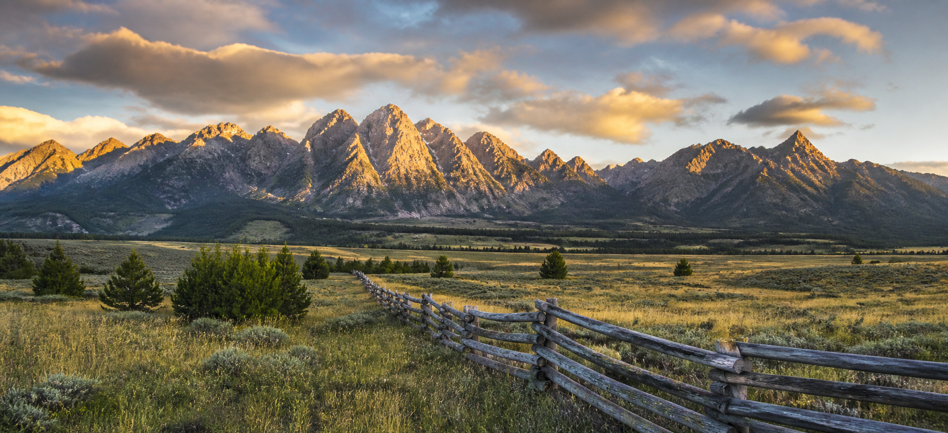 A split-rail fence running through a golden-hour valley toward a range of mountains catching the last light.